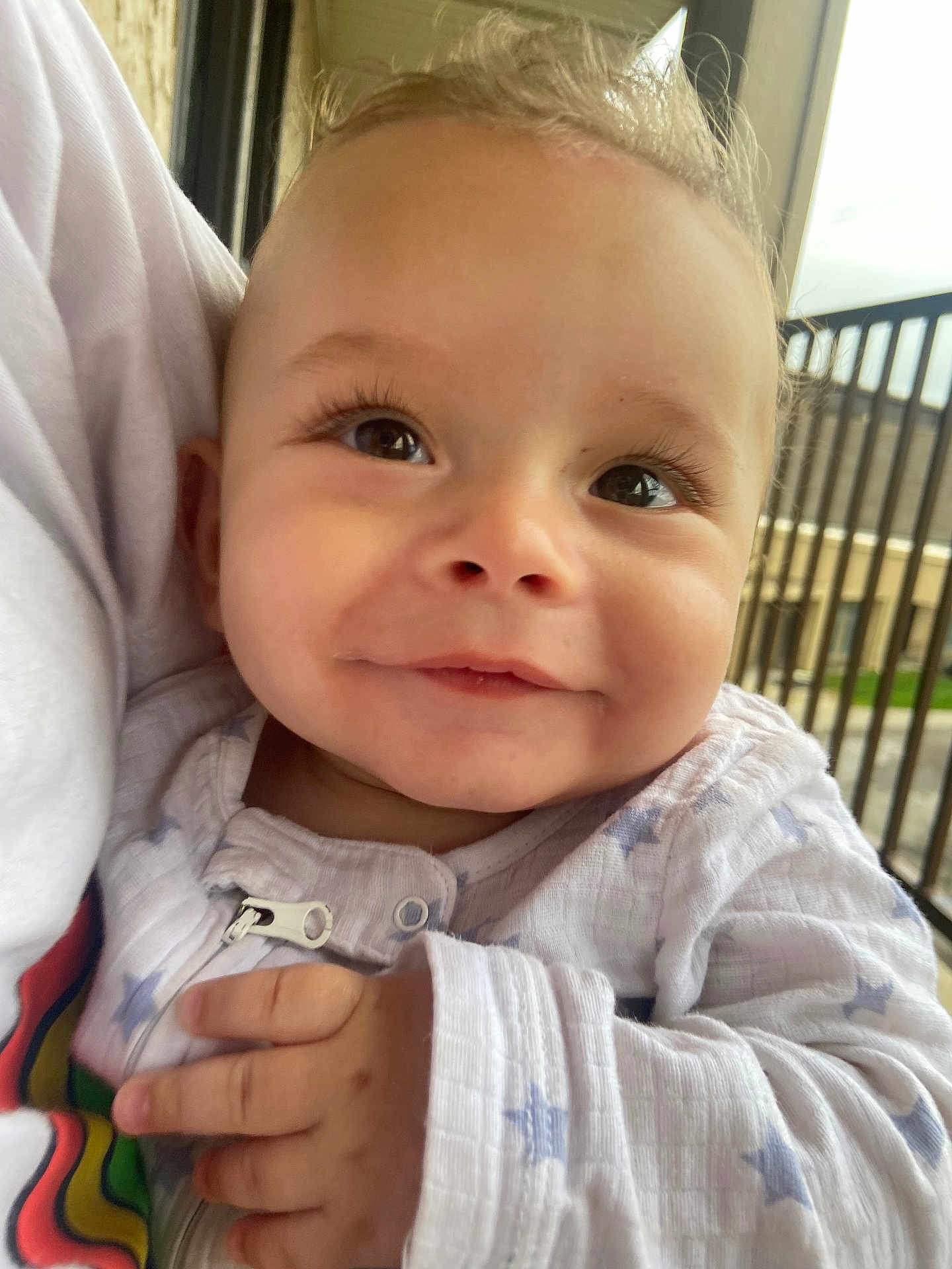 Camryn joined the competition — help win amazing prizes! baby, smiling, close_up, child, infant, outdoor, person, hand, clothing, stars_pattern, face, eyelashes, window, railing, building, daylight, holding, cute, portrait, happy