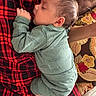 baby, sleeping, onesie, green_clothing, crocodile_print, person, red_plaid, lap, cushion, floral_pattern, indoor, cozy, resting, child, infant, peaceful, portrait, casual, comfort, home