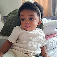 Eliora is registered to the contest to win money with this photo: baby, child, infant, headband, toy, bed, pillow, curly_hair, clothing, bracelet, indoor, portrait, cute, relaxed, soft_lighting, skin, hand, expression, white_clothes, seated