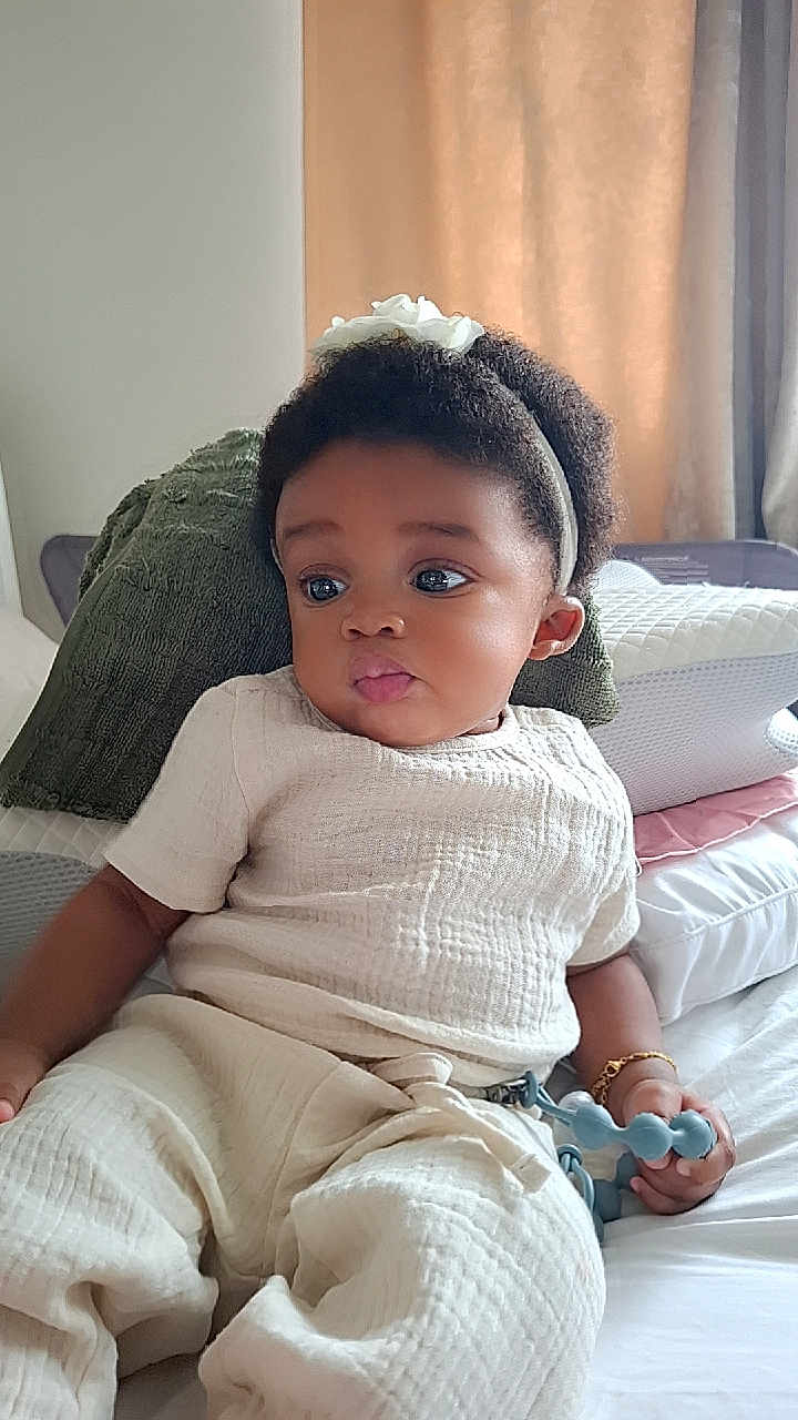 Eliora is registered to the contest to win money with this photo: baby, child, infant, headband, toy, bed, pillow, curly_hair, clothing, bracelet, indoor, portrait, cute, relaxed, soft_lighting, skin, hand, expression, white_clothes, seated