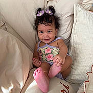 Khalessi is registered to the contest to win money with this photo: baby, child, clothing, cozy, cushion, cute, face, foot, hair_clips, hand, happy, indoors, infant, light_skin, pillow, portrait, purple_flowers, relaxed, sitting, smiling