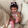 baby, child, smiling, hair_clips, purple_flowers, pillow, cushion, cute, infant, indoors, light_skin, happy, sitting, clothing, portrait, face, foot, hand, relaxed, cozy