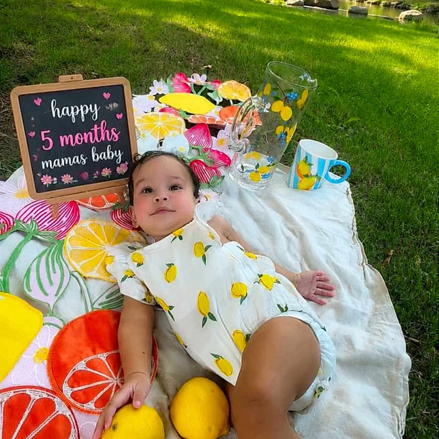 Khalessi is registered to the contest to win money with this photo: baby, blanket, chalkboard, child, clothing, cute, footwear, grass, infant, lemons, nature, outdoor, park, picnic, relaxing, sign, summer, sunlight, tree, willow_tree