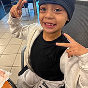 Koda joined the competition — help win amazing prizes! child, smile, peace_sign, beanie, black_shirt, gray_jacket, gray_pants, indoor, chair, table, floor_tiles, happy, casual_clothing, person, portrait, seated, light, young, cute, gesture