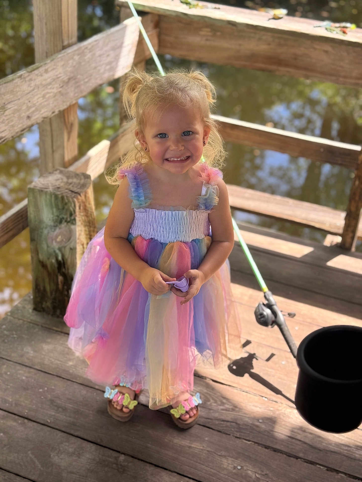 Paige is registered to the contest to win money with this photo: child, girl, smiling, dress, colorful, pigtails, outdoor, dock, wooden, water, fishing_rod, bucket, sunlight, happy, playful, summer, footwear, nature, portrait, cute