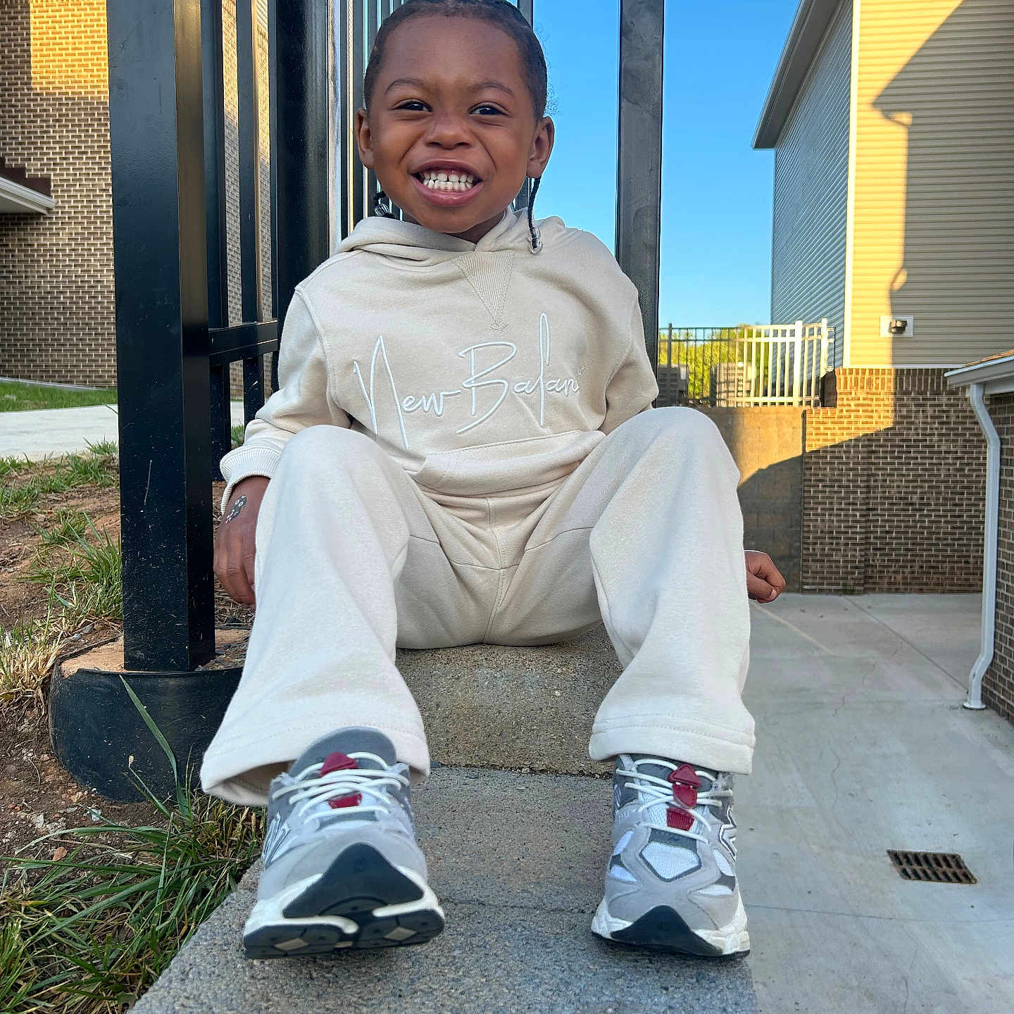Kaison is registered to the contest to win money with this photo: child, smiling, sitting, outdoor, concrete, sneakers, braids, hoodie, pants, daylight, building, fence, grass, sidewalk, happy, person, casual_clothing, urban, blue_sky, portrait