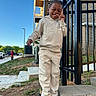 child, smiling, casual_clothing, hoodie, sweatpants, sneakers, outdoor, daylight, blue_sky, brick_building, metal_fence, steps, grass, sidewalk, urban, playful, gesture, happy, person, parked_cars