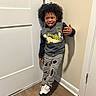 child, curly_hair, tongue_out, sweatshirt, simpsons, ripped_jeans, sneakers, door, wooden_floor, beige_wall, playful, standing, indoor, person, casual_clothing, happy, young, smiling, hand_gesture, portrait