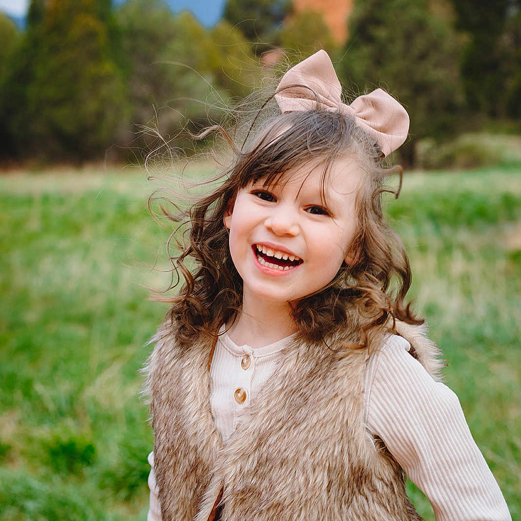Katie Gardner joined the competition — help win amazing prizes! bow, casual_clothing, child, curly_hair, cute, daylight, fur_vest, girl, grass, greenery, happy, joyful, long_sleeve_shirt, mountains, nature, outdoor, portrait, sitting, smiling, trees