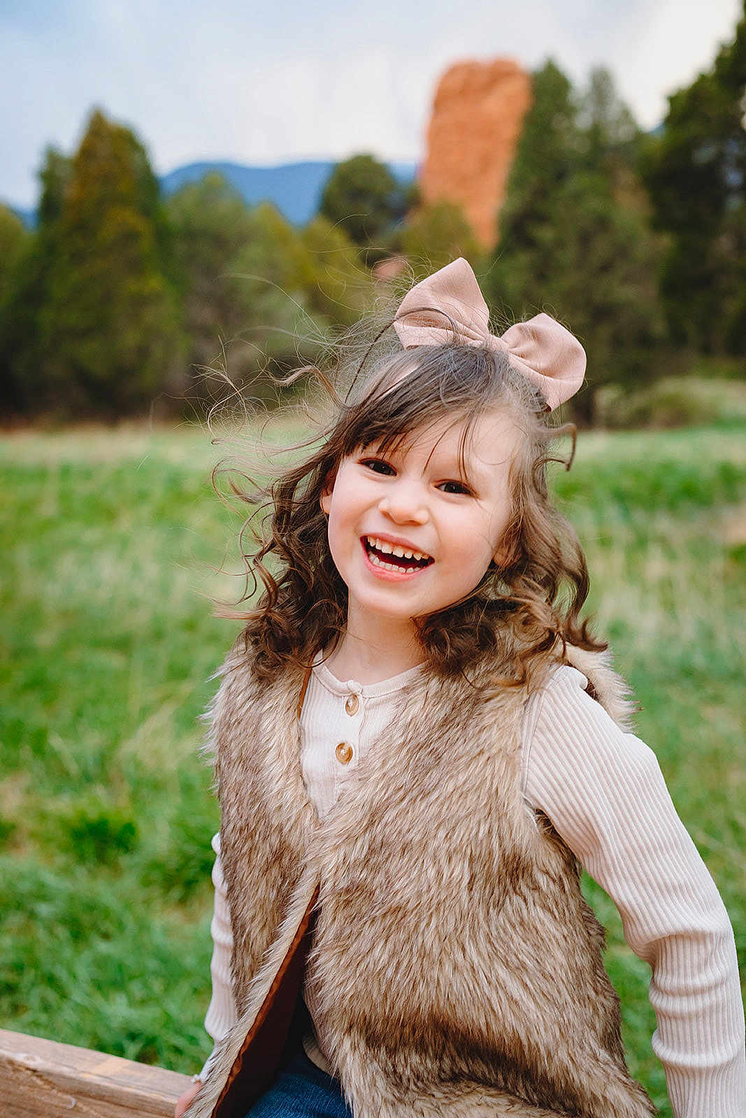 Katie Gardner joined the competition — help win amazing prizes! bow, casual_clothing, child, curly_hair, cute, daylight, fur_vest, girl, grass, greenery, happy, joyful, long_sleeve_shirt, mountains, nature, outdoor, portrait, sitting, smiling, trees