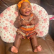 Egypt is registered to the contest to win money with this photo: baby, smiling, orange_headband, patterned_clothing, floral_cushion, brown_leather_couch, happy, infant, cute, sitting, indoors, portrait, chubby_cheeks, barefoot, headwear, child, young_child, home, comfort, cheerful