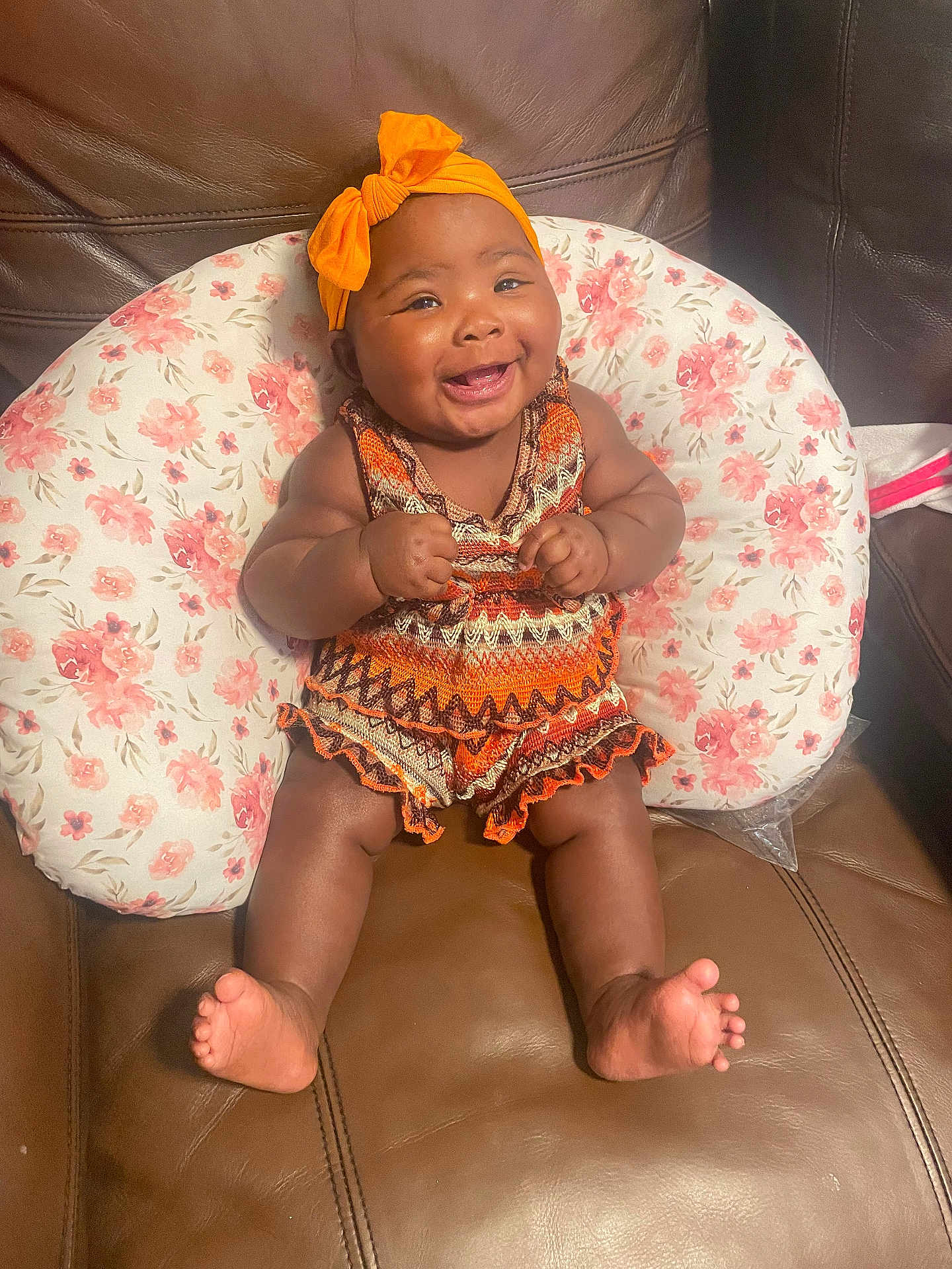 Egypt is registered to the contest to win money with this photo: baby, smiling, orange_headband, patterned_clothing, floral_cushion, brown_leather_couch, happy, infant, cute, sitting, indoors, portrait, chubby_cheeks, barefoot, headwear, child, young_child, home, comfort, cheerful