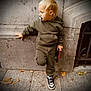 toddler, child, pacifier, hoodie, cargo_pants, sneakers, side_view, wall, stone_pavement, fallen_leaves, urban, outdoor, holding_object, casual_pose, blond_hair, young_child, street, footwear, curious, daylight