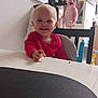 toddler, child, smiling, red_clothing, highchair, table, snack, indoor, home, photographs, toy, chair, face, happy, baby, person, furniture, light, background, cute