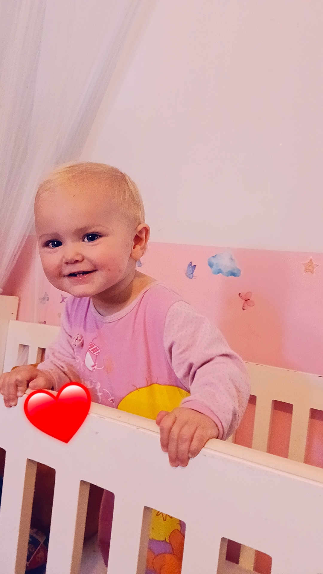 Hortense a rejoint le concours — aidez-le/la à gagner de superbes lots ! toddler, child, crib, smiling, blonde_hair, pink_wall, butterfly_stickers, cloud_sticker, heart_emoji, nursery, baby_clothes, hand, white_crib, cute, indoors, portrait, happy, young_child, room_decor, infant