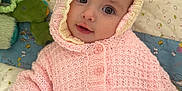 Nora is registered to the contest to win money with this photo: baby, child, knitted_hoodie, pink_clothing, quilt, soft_toys, indoor, portrait, cute, infant, smiling, buttoned_sweater, lying_down, cozy, warm_clothing, face, eyes, hand, fabric, patterned_background