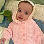 baby, child, knitted_hoodie, pink_clothing, quilt, soft_toys, indoor, portrait, cute, infant, smiling, buttoned_sweater, lying_down, cozy, warm_clothing, face, eyes, hand, fabric, patterned_background