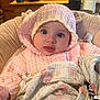 Nora joined the competition — help win amazing prizes! baby, infant, pink_clothing, knit_hat, blanket, indoors, face, wide_eyes, curious, cute, child, warm, cozy, seated, soft_texture, hoodie, home, kitchen, portrait, wrapped