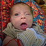 baby, infant, child, face, portrait, lying_down, blanket, patterned_blanket, striped_clothing, onesie, tongue, open_mouth, winking, blue_eyes, brown_hair, indoor, cozy, close_up, textile, humorous_expression