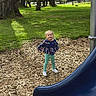 child, playground, slide, wood_chips, grass, trees, park, outdoor, daylight, casual_clothing, smiling, standing, young_child, nature, recreation, fun, blue_slide, greenery, happy, leisure