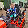 Lillian is registered to the contest to win money with this photo: child, girl, tractor, steering_wheel, outdoor, green_barn, blue_shirt, leggings, boots, dirt_ground, vehicle, farm_equipment, playful, person, seat, handle, farm, daylight, small_person, curly_hair