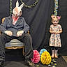 child, girl, person, costume, mask, bunny_mask, chair, candy, decorations, paper_eggs, boots, dress, ribbons, festive, background, seated, smile, indoor, dark_clothing, props