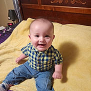 Harlynn joined the competition — help win amazing prizes! baby, child, smiling, sitting, indoor, bed, blanket, yellow_blanket, wooden_headboard, blue_jeans, checkered_shirt, happy, infant, person, face, sock, lamp, curtain, clock, furniture