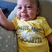 Nills a rejoint le concours — aidez-le/la à gagner de superbes lots ! baby, child, smiling, yellow_shirt, text_on_clothing, pillow, star_pattern, cushion, indoor, portrait, person, infant, cute, relaxed, casual_clothing, human, young_child, comfortable, happy, closeup