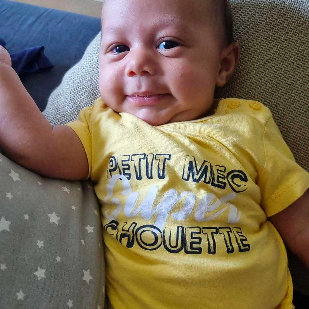 Nills a rejoint le concours — aidez-le/la à gagner de superbes lots ! baby, casual_clothing, child, closeup, comfortable, cushion, cute, happy, human, indoor, infant, person, pillow, portrait, relaxed, smiling, star_pattern, text_on_clothing, yellow_shirt, young_child