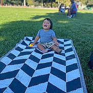 Zarel is registered to the contest to win money with this photo: toddler, child, blanket, grass, outdoor, park, toy, laughing, happy, person, summer, nature, greenery, casual_clothing, shorts, tshirt, barefoot, sunlight, trees, recreation