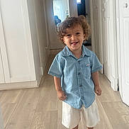 Liam is registered to the contest to win money with this photo: child, toddler, curly_hair, blue_shirt, white_shorts, sandals, hallway, door, floor, smile, standing, indoors, person, happy, light, wood_flooring, casual_clothing, portrait, cute, fashion