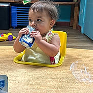 Cienna joined the competition — help win amazing prizes! baby, child, clothing, concentration, curly_hair, eating, face, floor, furniture, high_chair, indoor, plastic_container, room, shelf, sitting, snack_pouch, toddler, toys, wooden_table, yellow