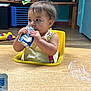 baby, child, clothing, concentration, curly_hair, eating, face, floor, furniture, high_chair, indoor, plastic_container, room, shelf, sitting, snack_pouch, toddler, toys, wooden_table, yellow