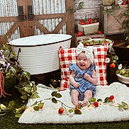 Paislee is registered to the contest to win money with this photo: baby, child, smiling, blue_outfit, white_bow, blanket, red_and_white_pillow, flowers, strawberries, basket, greenery, rustic, wooden_background, decor, fairy_lights, grass, indoor, cute, happy, portrait