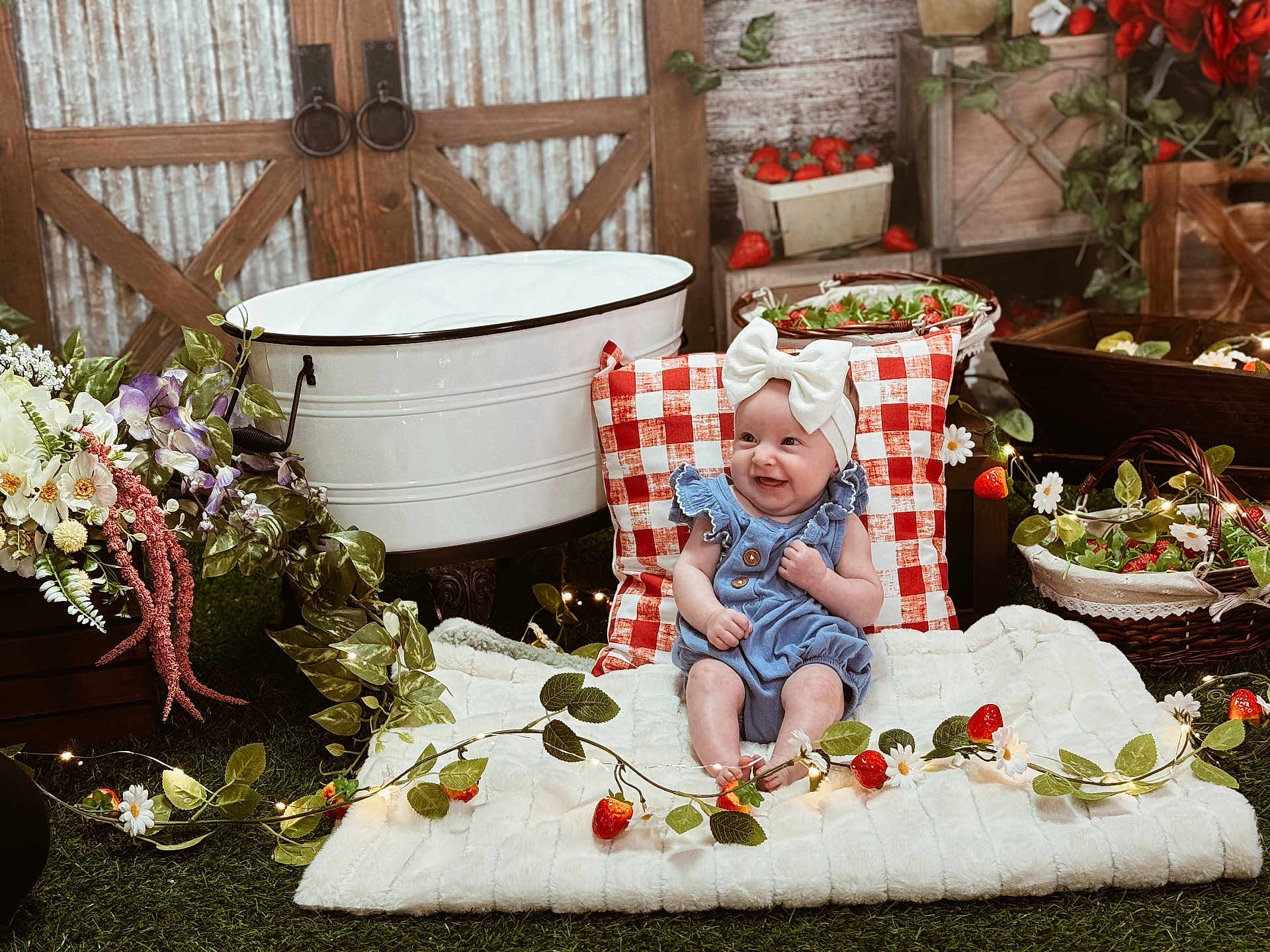 Paislee is registered to the contest to win money with this photo: baby, child, smiling, blue_outfit, white_bow, blanket, red_and_white_pillow, flowers, strawberries, basket, greenery, rustic, wooden_background, decor, fairy_lights, grass, indoor, cute, happy, portrait