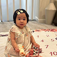 Melia joined the competition — help win amazing prizes! baby, child, girl, plush_toy, duck, dress, hair_clip, flower_pattern, blanket, carpet, indoor, sitting, cute, infant, soft_toy, foot, face, person, decor, cozy