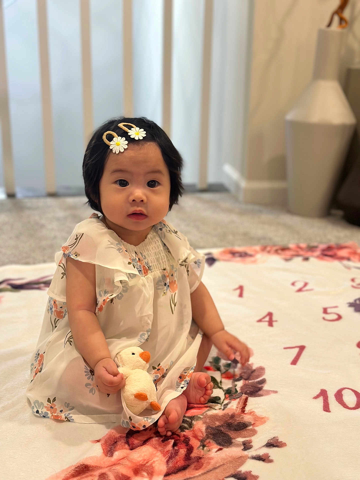 Melia joined the competition — help win amazing prizes! baby, child, girl, plush_toy, duck, dress, hair_clip, flower_pattern, blanket, carpet, indoor, sitting, cute, infant, soft_toy, foot, face, person, decor, cozy