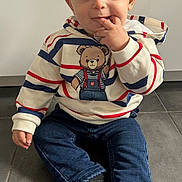 Milo a rejoint le concours — aidez-le/la à gagner de superbes lots ! toddler, child, indoor, floor, striped_hoodie, teddy_bear_graphic, jeans, black_socks, sitting, curious, fingers_in_mouth, cute, young_child, casual_clothing, portrait, person, playful, expression, tile_floor, young_kid