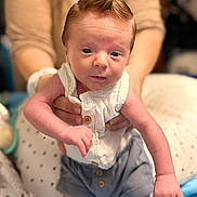 James joined the competition — help win amazing prizes! baby, infant, child, person, clothing, white_shirt, gray_shorts, hand, face, mohaawk_hairstyle, adult_hands, blurred_background, indoor, cute, newborn, skin, expression, portrait, sitting, holding
