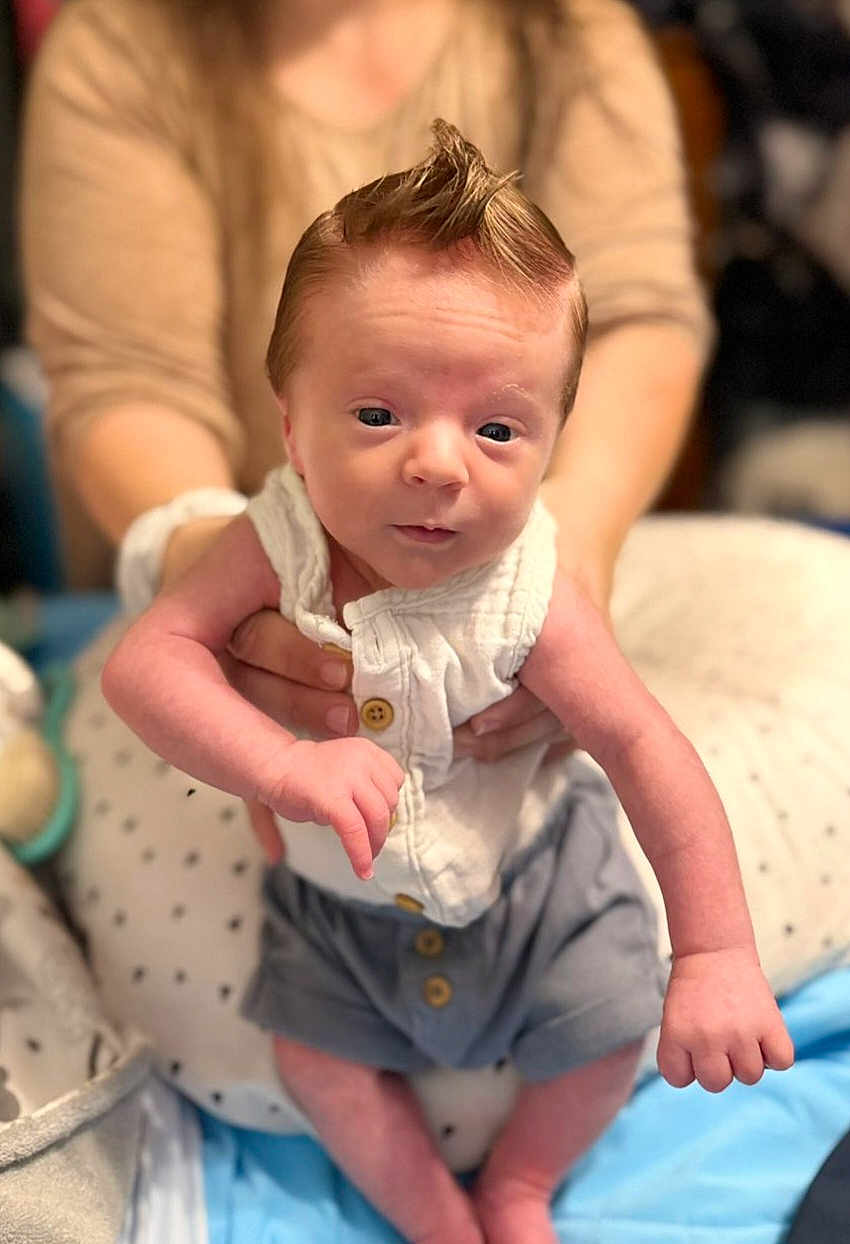 James joined the competition — help win amazing prizes! baby, infant, child, person, clothing, white_shirt, gray_shorts, hand, face, mohaawk_hairstyle, adult_hands, blurred_background, indoor, cute, newborn, skin, expression, portrait, sitting, holding