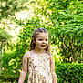 child, girl, floral_dress, outdoor, greenery, nature, smiling, happy, sunlight, summer, portrait, young, person, tree, grass, bush, daylight, casual, cute, playful