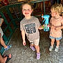 child, toddler, smiling, indoor, spray_bottle, fan, shorts, tshirt, footwear, curly_hair, toy, wooden_floor, green_railings, person, playful, casual_clothing, happy, cousin_crew, kids, family