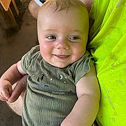Waylon is registered to the contest to win money with this photo: baby, smile, face, arm, green_clothing, pacifier, person, holding, indoor, infant, cute, happy, closeup, skin, child, toddler, short_sleeves, fabric, hand, person_holding