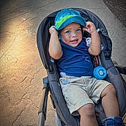 Legend is registered to the contest to win money with this photo: toddler, child, stroller, hat, blue_hat, smiling, outdoor, sidewalk, concrete, bottle, shorts, sneakers, blue_shirt, happy, baby, person, sunlight, shadow, casual_clothing, playful