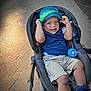 toddler, child, stroller, hat, blue_hat, smiling, outdoor, sidewalk, concrete, bottle, shorts, sneakers, blue_shirt, happy, baby, person, sunlight, shadow, casual_clothing, playful