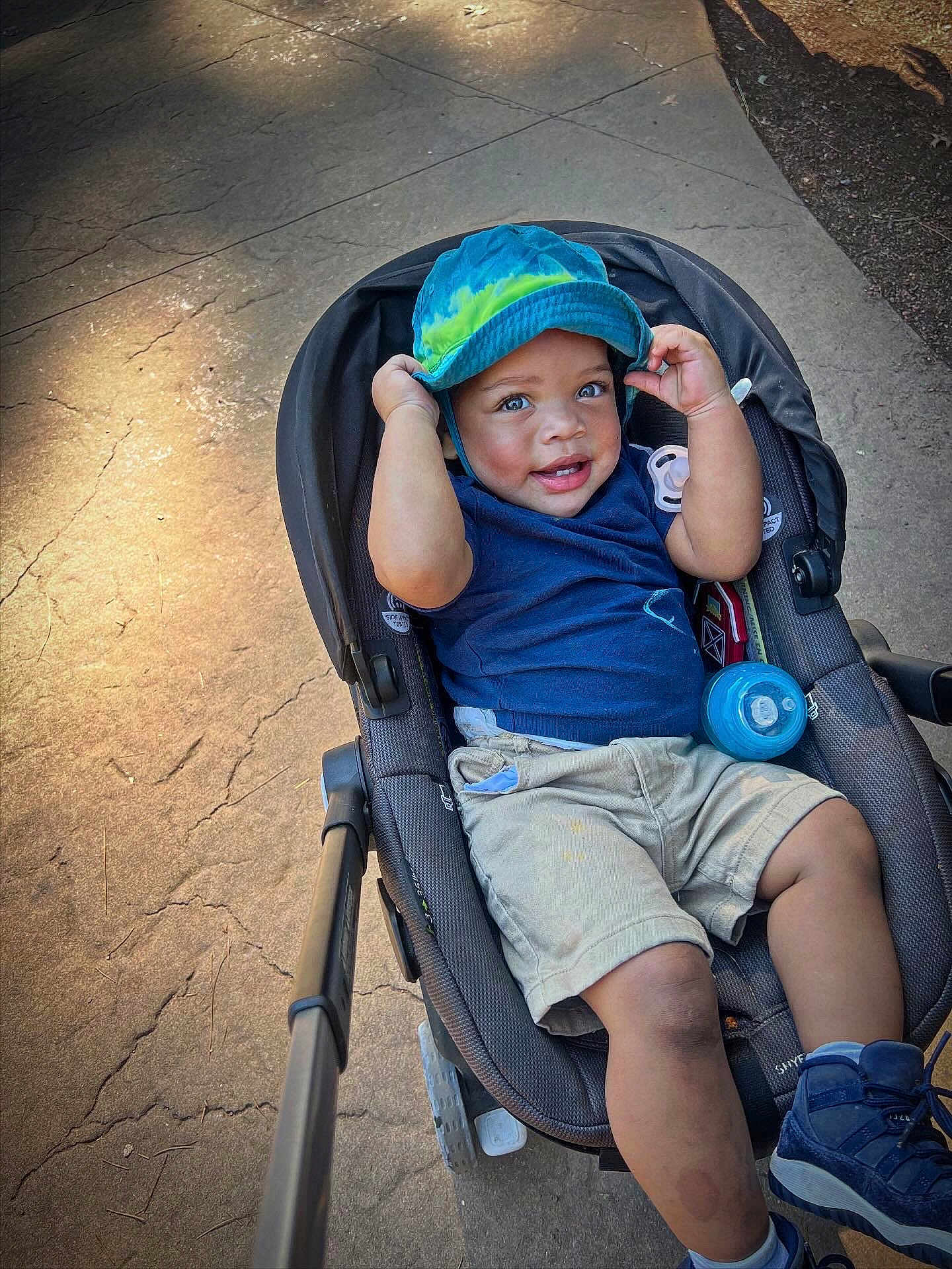 Legend is registered to the contest to win money with this photo: toddler, child, stroller, hat, blue_hat, smiling, outdoor, sidewalk, concrete, bottle, shorts, sneakers, blue_shirt, happy, baby, person, sunlight, shadow, casual_clothing, playful