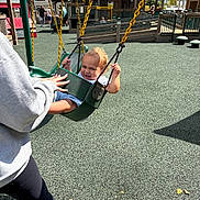 Armani is registered to the contest to win money with this photo: child, toddler, swing, playground, smiling, adult_hand, outdoor, daylight, curly_hair, safety_surface, play_equipment, casual_clothing, person, leisure, fun, happy, urban, recreation, park, family