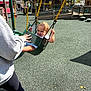 child, toddler, swing, playground, smiling, adult_hand, outdoor, daylight, curly_hair, safety_surface, play_equipment, casual_clothing, person, leisure, fun, happy, urban, recreation, park, family