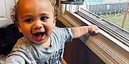 Armani is registered to the contest to win money with this photo: toddler, child, smiling, curly_hair, indoor, window, shirt, reaching_hand, happy, daylight, jacket, bedroom, carpet, home, person, furniture, playful, young_child, expression, casual_clothing