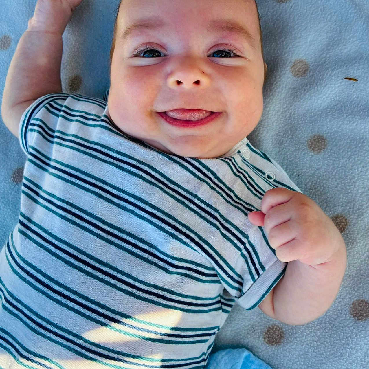 Dilan is registered to the contest to win money with this photo: baby, blanket, child, clothing, cute, daylight, eyes, face, hand, happy, head, infant, lying_down, outdoor, polka_dots, skin, smiling, soft, striped_shirt, tongue_out